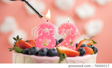 Birthday cake number 82, pink candle on beautiful cake with berries and lighter with fire against background of white clouds and pink sky. Close-up 78089317