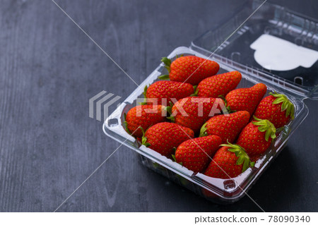 Picture of a box of fresh red strawberry. The strawberry come with green leaves. The box is a clear plastic box. It is placed on a black wooden table. 78090340