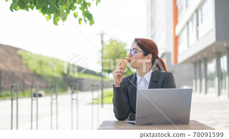 A smiling woman works remotely on a laptop and enjoys eating an ice cream cone while sitting at a wooden table. Portrait of a girl in a business suit for a break in a street cafe on a hot summer day. A smiling woman works remotely on a laptop and enjoys eating an ice cream cone while sitting at a wooden table. Portrait of a girl in a business suit for a break in a street cafe on a hot summer day. 78091599