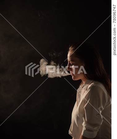 Pensive brunette in a white shirt smokes a hookah in the dark. Portrait of a woman smoking a vape, hovering. Black background. Electronic Cigarette. Bad habit. Pensive brunette in a white shirt smokes a hookah in the dark. Portrait of a woman smoking a vape, hovering. Black background. Electronic Cigarette. Bad habit. 78096747