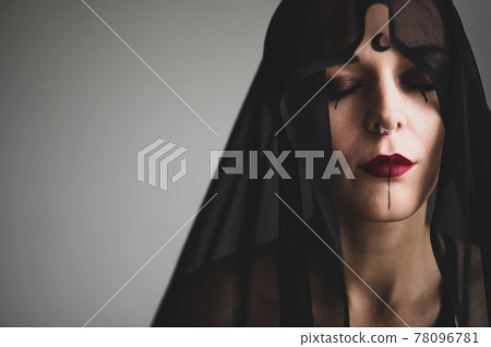 A grinning woman in a black veil with dark makeup and an earring in her nose. Halloween Dead Bride Costume. Portrait of the sinister Witch in front of the coven. 78096781