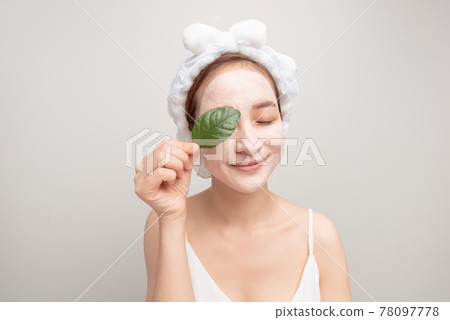 Beauty portrait of woman in towel on head with white nourishing mask or creme on face and green leaf in hand, 78097778
