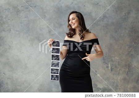 Pregnant woman pointing at her tummy and showing ultrasound scan image on gray 78100088