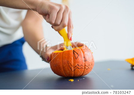 A woman cuts out a face with a stationery knife on a pumpkin for Halloween lamps on a black table. Close-up of female hands making jack-o'-lantern on the eve of all saints. Front view. 78103247