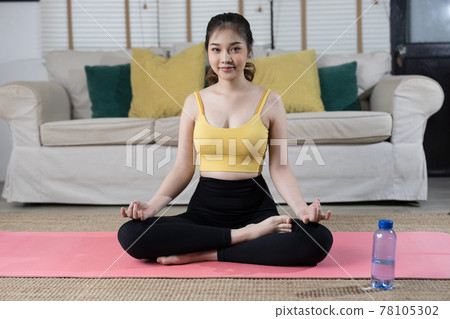 Asian woman practicing yoga in her home. She is wearing yoga outfit while sitting on the yoga mat. 78105302