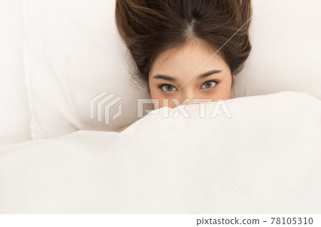 Young asian woman laying on her bed. She can be seen covering her mouth under the blanket and only showing her eye Young asian woman laying on her bed. She can be seen covering her mouth under the blanket and only showing her eye 78105310