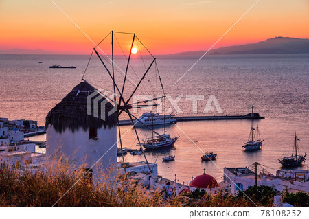 Famous traditional Greek windmill over port of Mykonos, Greece at sunset. Beautiful sky, sun touch sea horizon, boats, pier, high grass in foreground. 78108252