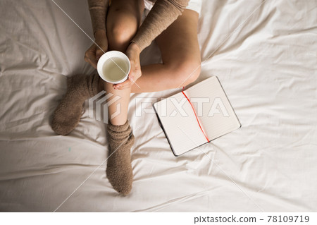 Top view of woman sitting on her bed with her notebook open beside her. She is also holding a cup of water in her hand. She wear wool sock. Top view of woman sitting on her bed with her notebook open beside her. She is also holding a cup of water in her hand. She wear wool sock. 78109719
