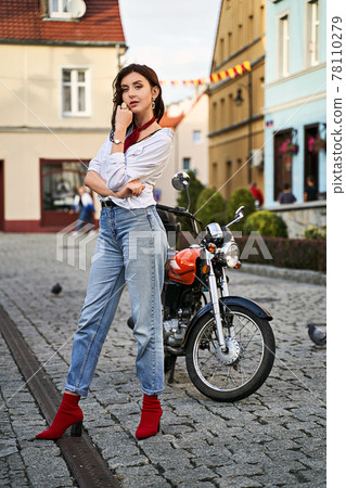 Young beautiful woman in red knitted fabric block heel sock shoes, blue denim jeans pants and teddy jacket coat posing near a motorcycle on a street Young beautiful woman in red knitted fabric block heel sock shoes, blue denim jeans pants and teddy jacket coat posing near a motorcycle on a street 78110279