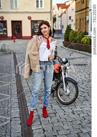 Young beautiful woman in red knitted fabric block heel sock shoes, blue denim jeans pants and teddy jacket coat posing near a motorcycle on a street 78110280