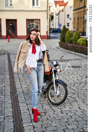 Young beautiful woman in red knitted fabric block heel sock shoes, blue denim jeans pants and teddy jacket coat posing near a motorcycle on a street Young beautiful woman in red knitted fabric block heel sock shoes, blue denim jeans pants and teddy jacket coat posing near a motorcycle on a street 78110281
