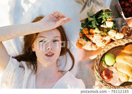 Top view candid portrait of young red-haired woman lying on a picnic blanket near flowers and basket of food. Eco-friendly lifestyle, Summer Vacation, relaxation, summer Picnic Top view candid portrait of young red-haired woman lying on a picnic blanket near flowers and basket of food. Eco-friendly lifestyle, Summer Vacation, relaxation, summer Picnic 78112185