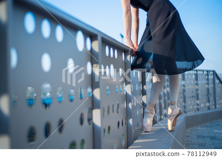 Ballerina in ballet legs in shoes and black tutu dancing by the fence. Beautiful young woman in black dress and pointe dancing outside. Gorgeous ballerina performing a dance outdoors. Close up 78112949