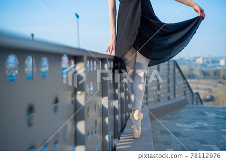 Ballerina in ballet legs in shoes and black tutu dancing by the fence. Beautiful young woman in black dress and pointe dancing outside. Gorgeous ballerina performing a dance outdoors. Close up 78112976