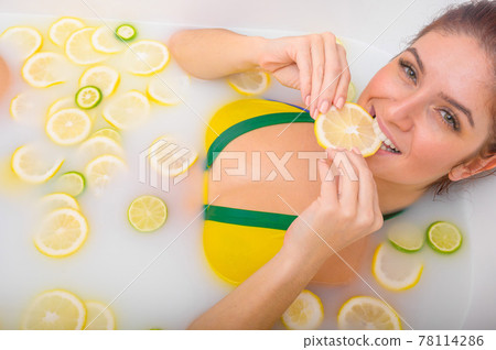 Beautiful young red-haired woman takes a bath with milk and citrus fruits. Portrait of Caucasian smiling girl eating lemon wedge in skin whitening bath. Taking care of the body. 78114286
