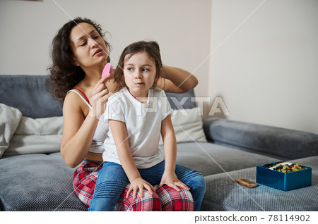 Adorable girl sitting on her mother's legs while she is combing her hair Adorable girl sitting on her mother's legs while she is combing her hair 78114902