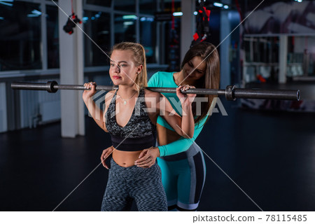 Young athletic girl doing barbell squats in the gym under the supervision of a trainer. A female coach teaches a ward to squat with a bar. Personal training. Exercises on the buttocks. Young athletic girl doing barbell squats in the gym under the supervision of a trainer. A female coach teaches a ward to squat with a bar. Personal training. Exercises on the buttocks. 78115485