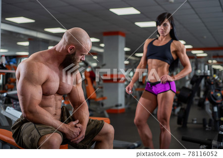 A young muscular guy is sitting on a bench in the gym and resting after a workout. Behind is a muscular dark-haired girl in shorts. Fitness center in the gym. A young muscular guy is sitting on a bench in the gym and resting after a workout. Behind is a muscular dark-haired girl in shorts. Fitness center in the gym. 78116052