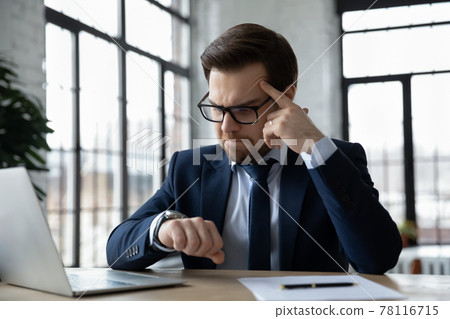Concerned businessman sitting at workplace and consulting wrist watch Concerned businessman sitting at workplace and consulting wrist watch 78116715