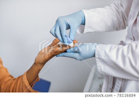 Close up cropped photo of hands of a female lab worker in white coat and gloves, taking a patient's blood sample, using painless scarifier. Black woman patient ready to blood test. Close up cropped photo of hands of a female lab worker in white coat and gloves, taking a patient's blood sample, using painless scarifier. Black woman patient ready to blood test. 78116884