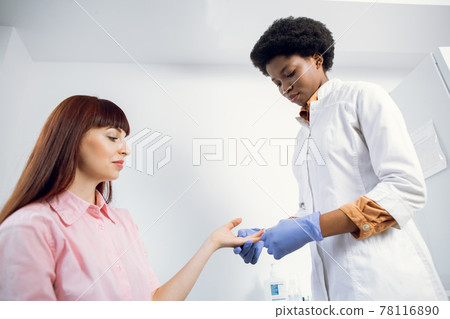 Concentrated young afro american woman doctor doing blood collecting analysis for her female Caucasian patient in modern hospital. Blood analysis, group test, HIV, hepatitis. Concentrated young afro american woman doctor doing blood collecting analysis for her female Caucasian patient in modern hospital. Blood analysis, group test, HIV, hepatitis. 78116890