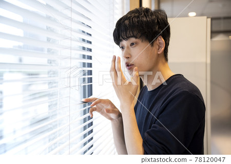 A young man who is surprised to look out from the window blinds A young man who is surprised to look out from the window blinds 78120047