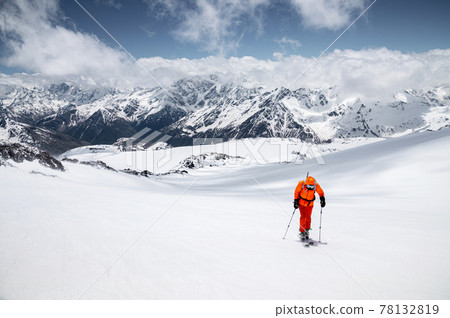 Young male freerider guide in a yellow ski suit with a backpack on his shoulders climbing the mountain on skis on a sunny day. 78132819