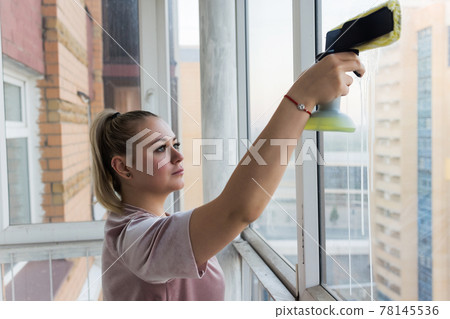 Beautiful young housewife in working clothes washing windows by professional tools at home. Beautiful young housewife in working clothes washing windows by professional tools at home. 78145536