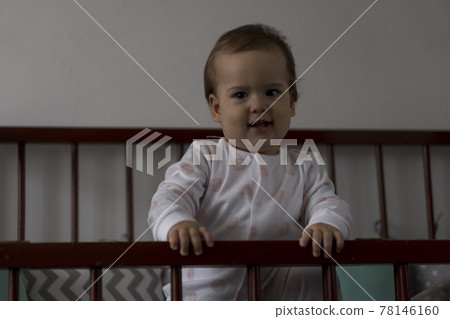 Cute cheerful little chubby baby girl sitting in grey baby crib after waking up from sleep in white and pink pajamas with teddy bear and smiling. Childhood, leisure, comfort, medicine, health concept 78146160