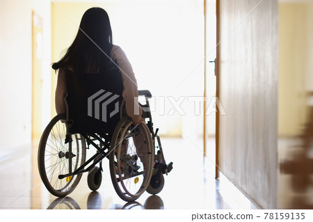 Woman in wheelchair in dark hallway looks out window Woman in wheelchair in dark hallway looks out window 78159135