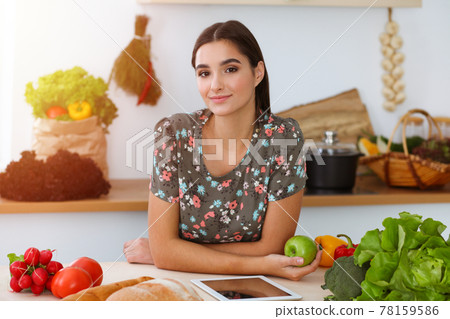 An attractive young dark-haired woman holding green apple and choosing the recipe for a delicious meal while smiling in sunny kitchen. Tablet pc is the best cookbook An attractive young dark-haired woman holding green apple and choosing the recipe for a delicious meal while smiling in sunny kitchen. Tablet pc is the best cookbook 78159586
