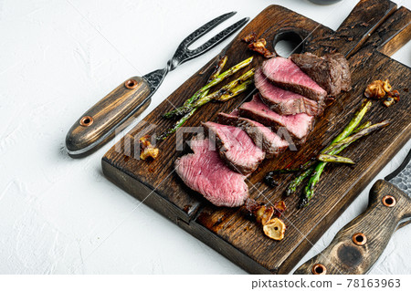 Fillet Mignon tenderloin Grilled and sliced meat beef steaks medium rare, with onion and asparagus, on wooden serving board, with meat knife and fork, on white stone background, with copy space for 78163963