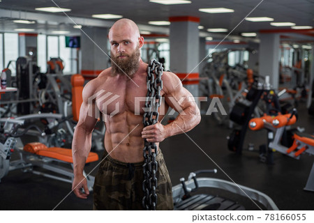 A bald, bearded bodybuilder with large relief muscles holds a heavy thick chain in his hands. Bright gym. 78166055