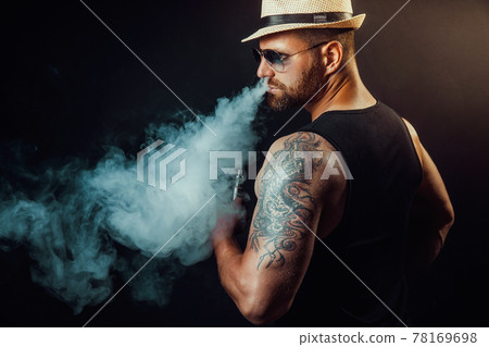 Bearded brutal male in hat and sunglasses smoking a vapor cigarette as an alternative to tobacco. Studio shot on dark background. 78169698