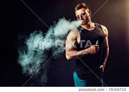 Bearded brutal male in sunglasses smoking a vapor cigarette as an alternative to tobacco. Studio shot on dark background. 78169718