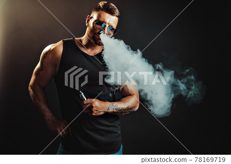Bearded brutal male in sunglasses smoking a vapor cigarette as an alternative to tobacco. Studio shot on dark background. 78169719