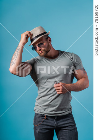 Bearded male in hat and sunglasses smoking a vapor cigarette as an alternative to tobacco. Studio shot on blue background. 78169720