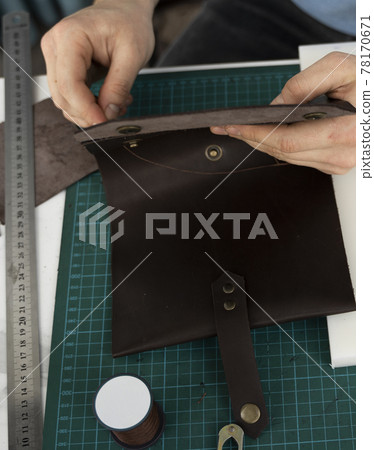 Mens hand working on the leather wallet in his workshop. Working process with a brown natural leather. Craftsman holding a crafting tools. 78170671