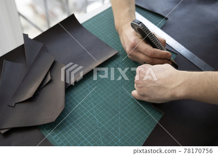 Men s hand holding a stationery knife and metal ruler and cutting on a pieces for a leather wallet in his workshop. Working process with a brown natural leather. Craftsman holding a crafting tools. 78170756