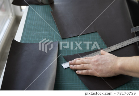 Men s hand holding a stationery knife and metal ruler and cutting on a pieces for a leather wallet in his workshop. Working process with a brown natural leather. Craftsman holding a crafting tools. 78170759