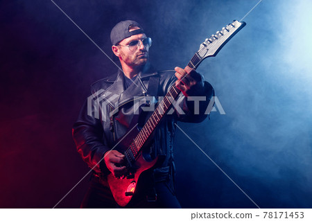 Brutal bearded Heavy metal musician in leather jacket and sunglasses is playing electrical guitar. Shot in a studio on dark background with smoke Brutal bearded Heavy metal musician in leather jacket and sunglasses is playing electrical guitar. Shot in a studio on dark background with smoke 78171453