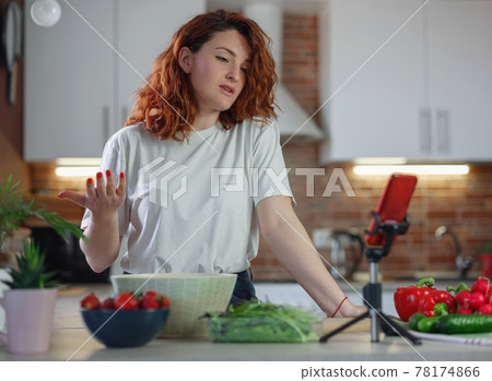 Young pretty woman blogger shoots a video of a salad recipe on a smartphone camera. Young pretty woman blogger shoots a video of a salad recipe on a smartphone camera. 78174866