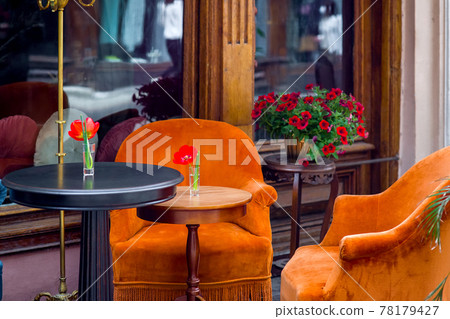 2 round wooden tables in black and brown with soft orange armchairs at front of building with large wooden window frames and stand for flowerpot with blooming petunias, outdoor cafe with empty seats. 2 round wooden tables in black and brown with soft orange armchairs at front of building with large wooden window frames and stand for flowerpot with blooming petunias, outdoor cafe with empty seats. 78179427