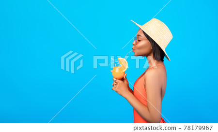 Side view of joyful black lady in swimwear having beach vacation, sipping tropical cocktail, blue background 78179967