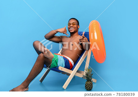 Flirty African American guy in swimwear showing CALL ME gesture, pointing at camera, sitting in lounge chair Flirty African American guy in swimwear showing CALL ME gesture, pointing at camera, sitting in lounge chair 78180039