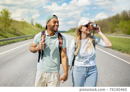 Happy young multiracial couple hitchhiking together, holding hands, taking photos on camera, walking along road Happy young multiracial couple hitchhiking together, holding hands, taking photos on camera, walking along road 78180460