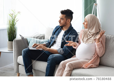 Annoyed pregnant muslim woman looking at husband that spending time with laptop Annoyed pregnant muslim woman looking at husband that spending time with laptop 78180488