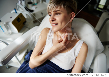 Close-up of seductive young blonde woman massaging her neck. Performing lymphatic drainage massage using a roller massager with jade stones. Close-up of seductive young blonde woman massaging her neck. Performing lymphatic drainage massage using a roller massager with jade stones. 78181623