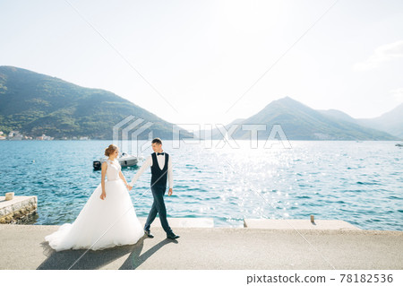 The bride and groom are walking hand in hand on the road by the sea, behind them are mountains and a boat The bride and groom are walking hand in hand on the road by the sea, behind them are mountains and a boat 78182536