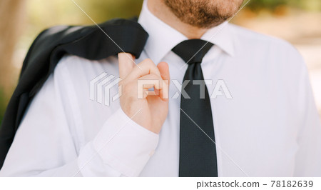 A man in white shirt and black tie stands and holds a jacket over his shoulder, close-up A man in white shirt and black tie stands and holds a jacket over his shoulder, close-up 78182639